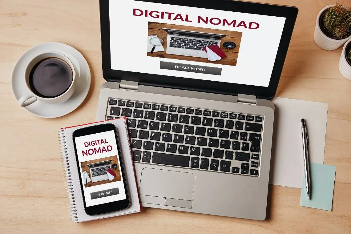 The digital nomad work table contains the necessary equipment, a coffee mug, laptop, sticky notes, gardening pots, and pens, and both computer and mobile screen has the word, digital nomad.